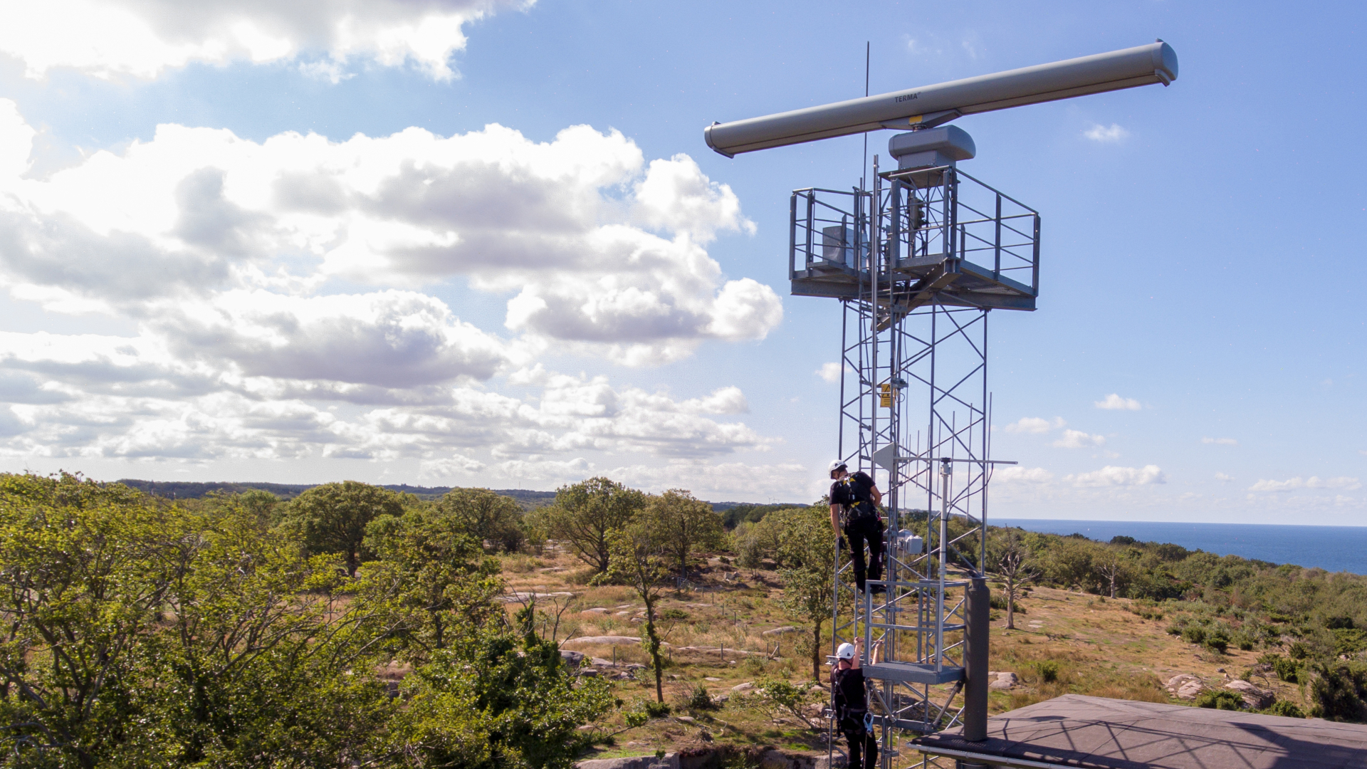 To mænd klatrer op ad en stige på en mast med en stor radar siddende på toppen. Buskads og skyer i baggrunden.