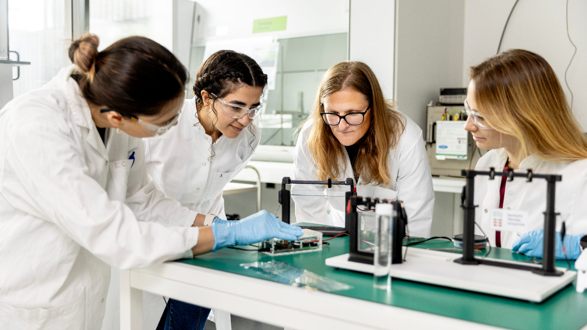 Fire kvinder i hvide kitler står i et laboratorie og kigger på nogle instrumenter.